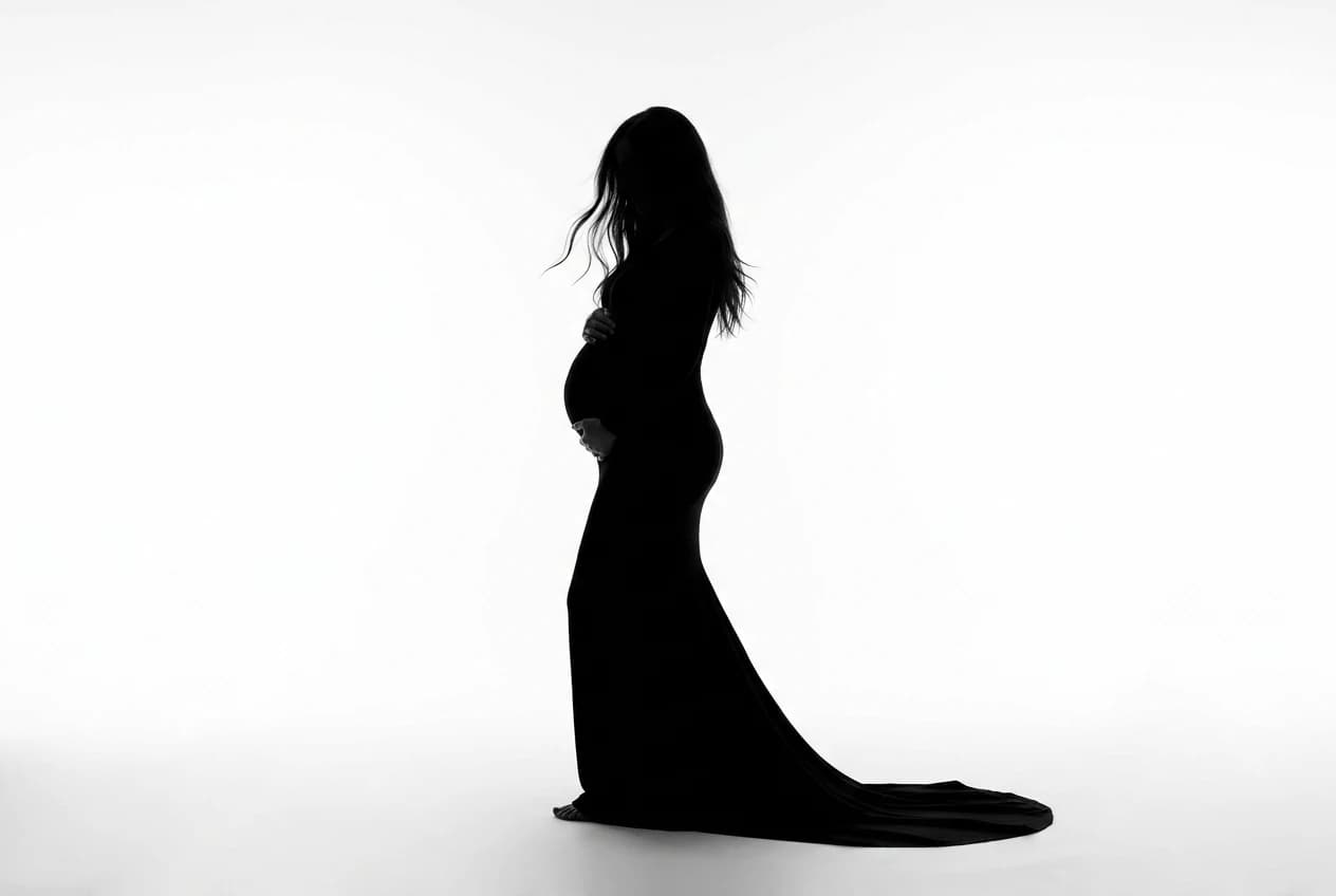 Pure black silhouette of a pregnant woman in a flowing floor-length gown against a high-key white studio backdrop, hands cradling her belly — the editorial register this system was built to book