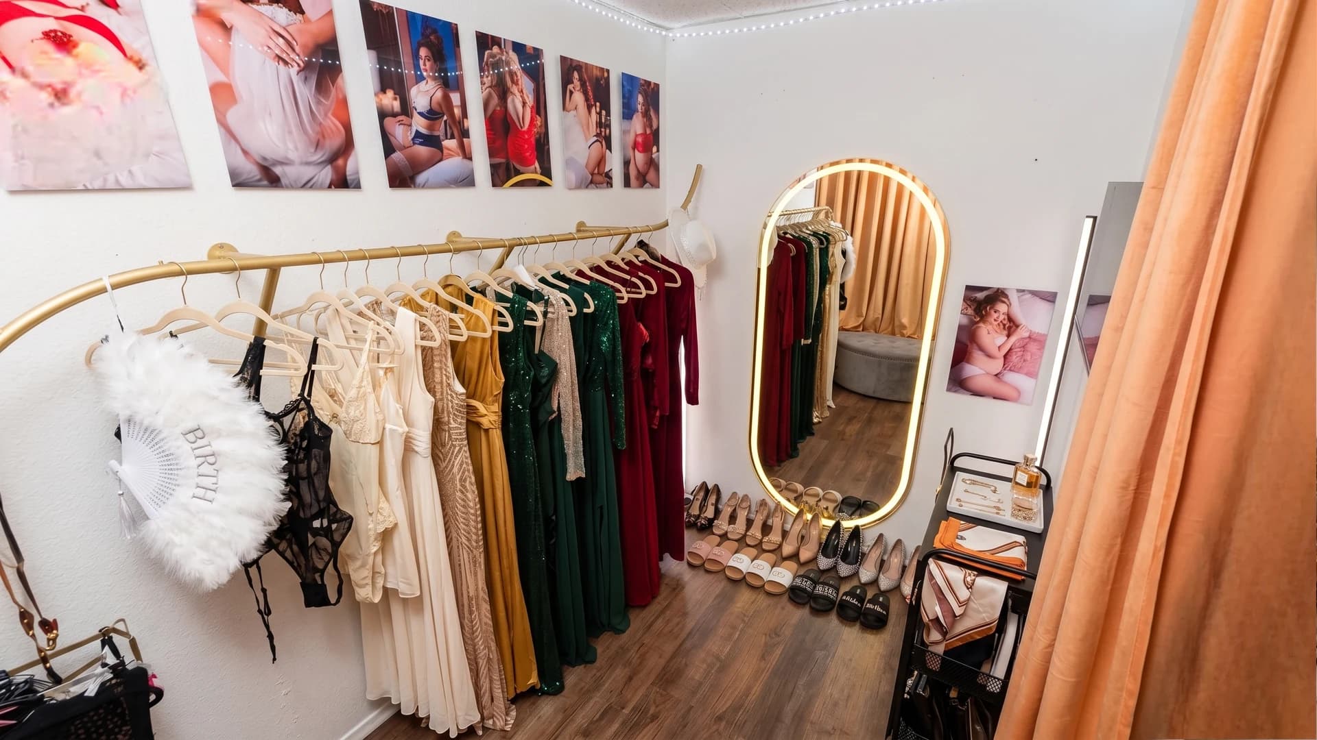 Casey Quist's studio wardrobe room — gold garment rack with gowns and lingerie, oval lit mirror, shoes lined up on the floor, boudoir prints on the wall